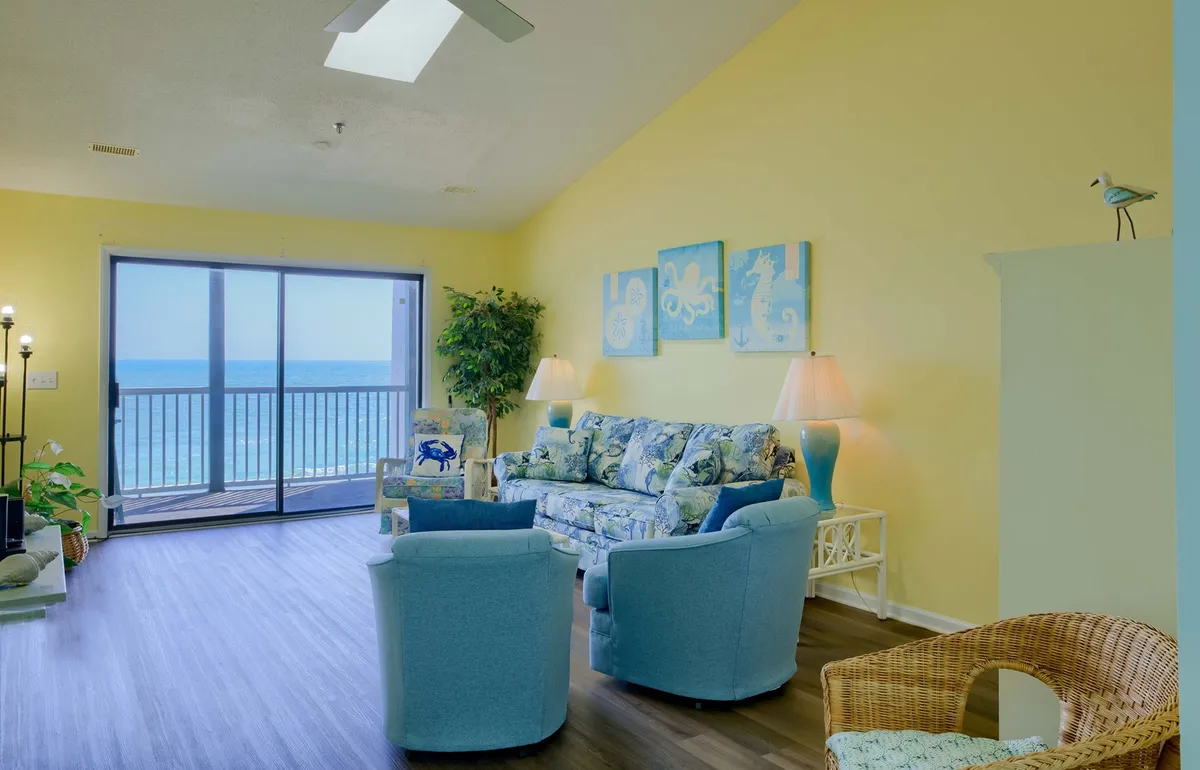 This image features a bright, airy living area with large sliding glass doors that open up to an inviting ocean view. The space is decorated with a blue and white couch, blue accent chairs, and a tropical plant, all bathed in cheerful, natural light. The ocean can be glimpsed just beyond, giving a sense of direct beachfront access.