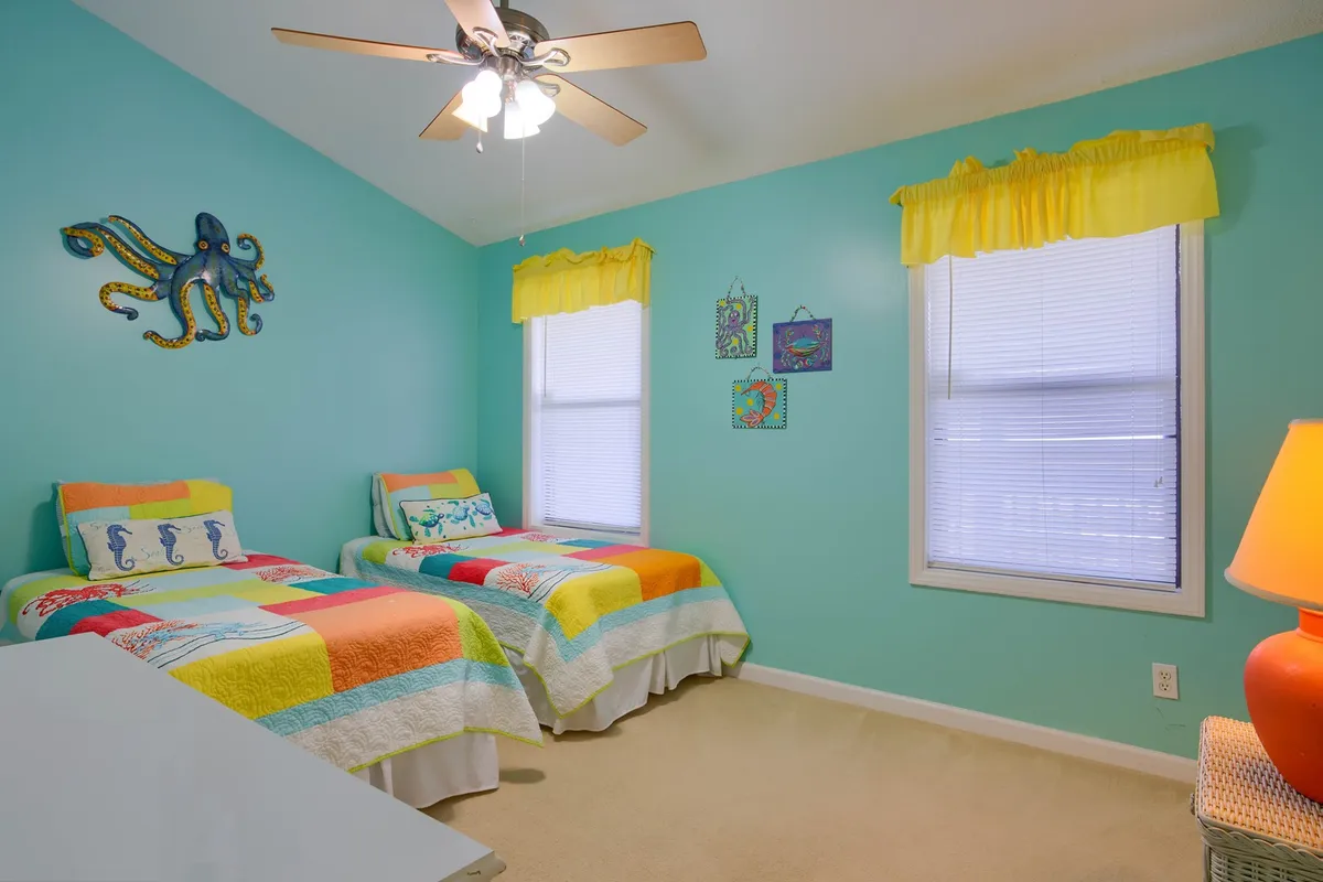 A cheerful twin bedroom featuring two beds with colorful quilts, octopus wall art, vibrant yellow curtains, and a playful table lamp. The room is light-filled, creating a fun and welcoming space suited for children or friends.