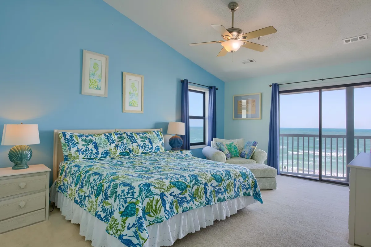 A serene bedroom featuring a king-sized bed with colorful bedding, positioned to take advantage of large windows that offer a sweeping view of the ocean. A comfortable armchair is set by the window, inviting relaxation and moments of quiet as sunlight pours in.