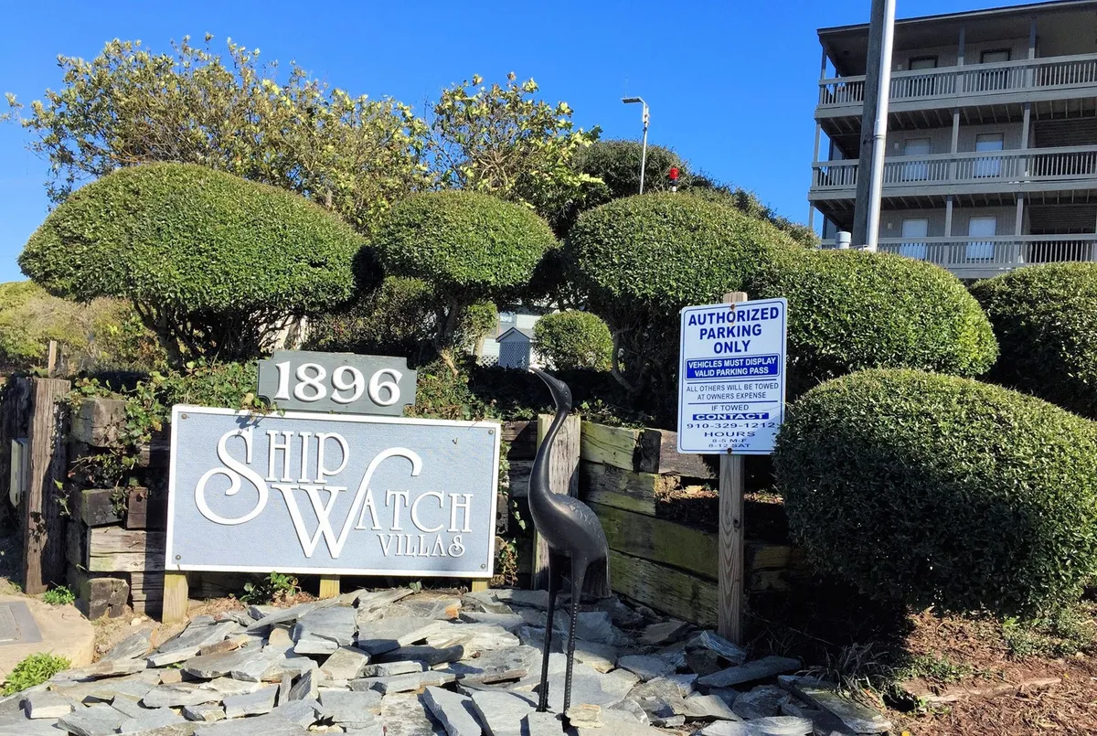 An exterior shot of the Ship Watch Villas sign accented by manicured shrubs, a decorative bird sculpture, and visible parking signage. The parking area is inviting and well-marked, providing clarity and peace of mind for guests arriving by car.