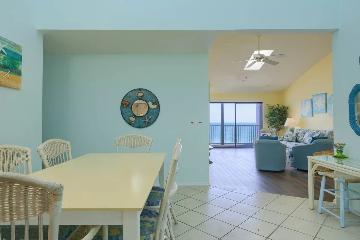 A cheerful and inviting open-plan space where the dining table sits adjacent to a comfortable blue couch, all set against a backdrop of expansive ocean views visible through large windows. Tasteful wall art and coastal decor reinforce the relaxed seaside atmosphere.