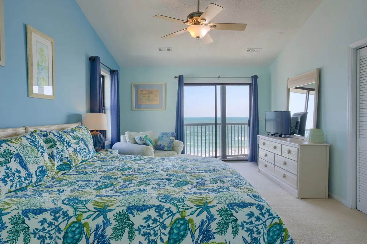A serene bedroom with a bed featuring floral bedding, large windows offering stunning ocean views, a cozy armchair, and a dresser topped with a TV. Ample natural light suggests a direct connection to a balcony overlooking the Atlantic.