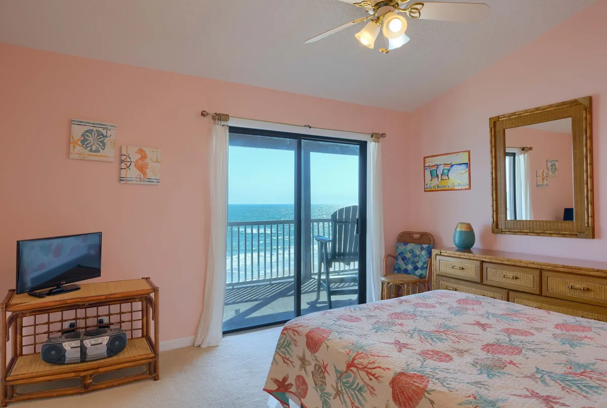 A peaceful bedroom with a coral-patterned blanket on the bed, a TV on the dresser, and a private balcony accessed through sliding doors. The balcony offers stunning ocean views, and the room is filled with natural light reflected by a large mirror.