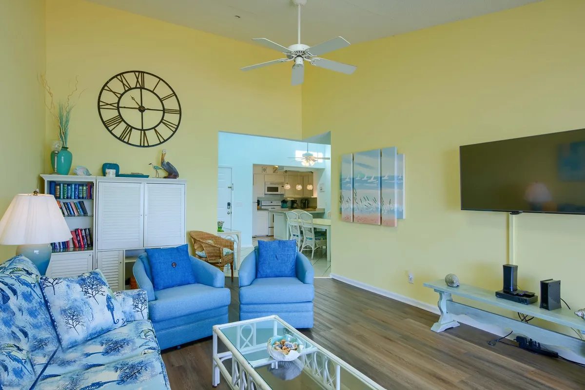 This image features a bright and inviting living room with a blue floral sofa and matching accent chairs, a large wall clock, and beach-themed artwork. The space is open and arranged for group conversation, with a coffee table central to family game nights or relaxed gatherings. Sunlight pours in, suggesting proximity to large windows or a balcony.