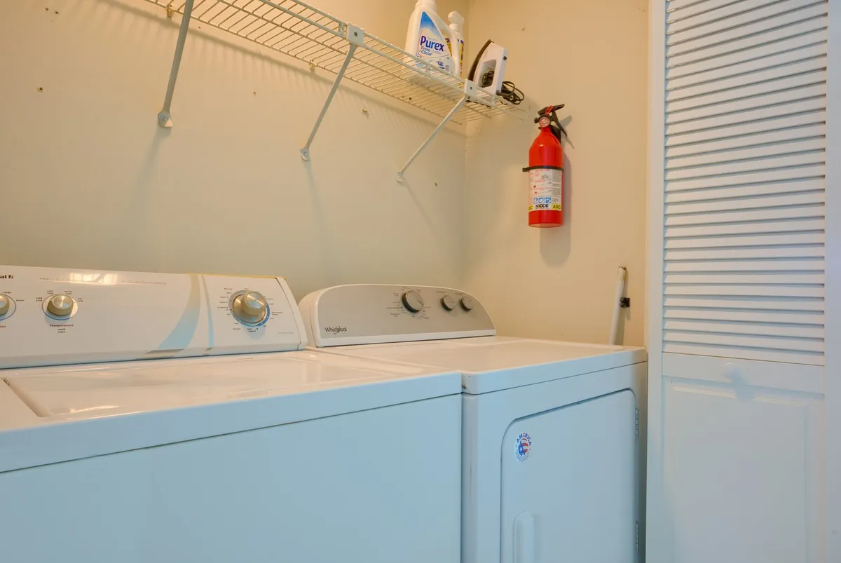 A compact laundry closet featuring a washing machine and dryer neatly stacked side by side, with a shelf above stocked with laundry essentials and a fire extinguisher for safety. The setup is clean and practical, emphasizing easy in-unit laundry access.