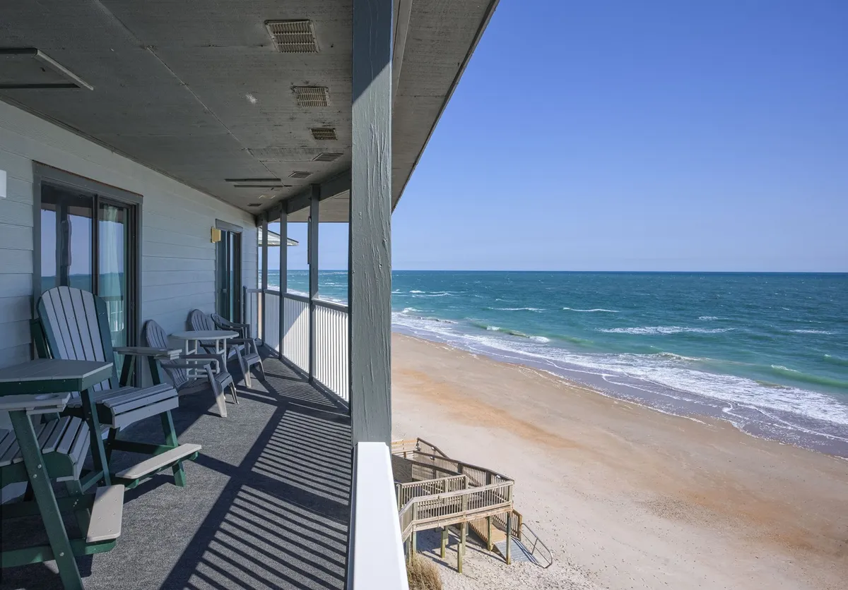 The image shows a private balcony at Shipwatch Villas overlooking the pristine sands of North Topsail Beach, with comfortable chairs positioned to take in the sweeping Atlantic Ocean views. Sunlight sparkles on the water, casting a peaceful glow, and the beachfront stretches invitingly below, perfect for morning coffee or evening relaxation.