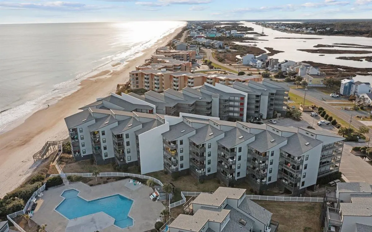 A wide-angle view of the Shipwatch Villas' oceanfront swimming pool, with sparkling water surrounded by a sun deck and sun loungers. The scene includes the beachfront just beyond, with gentle waves lapping the shore and the villa buildings in the background.