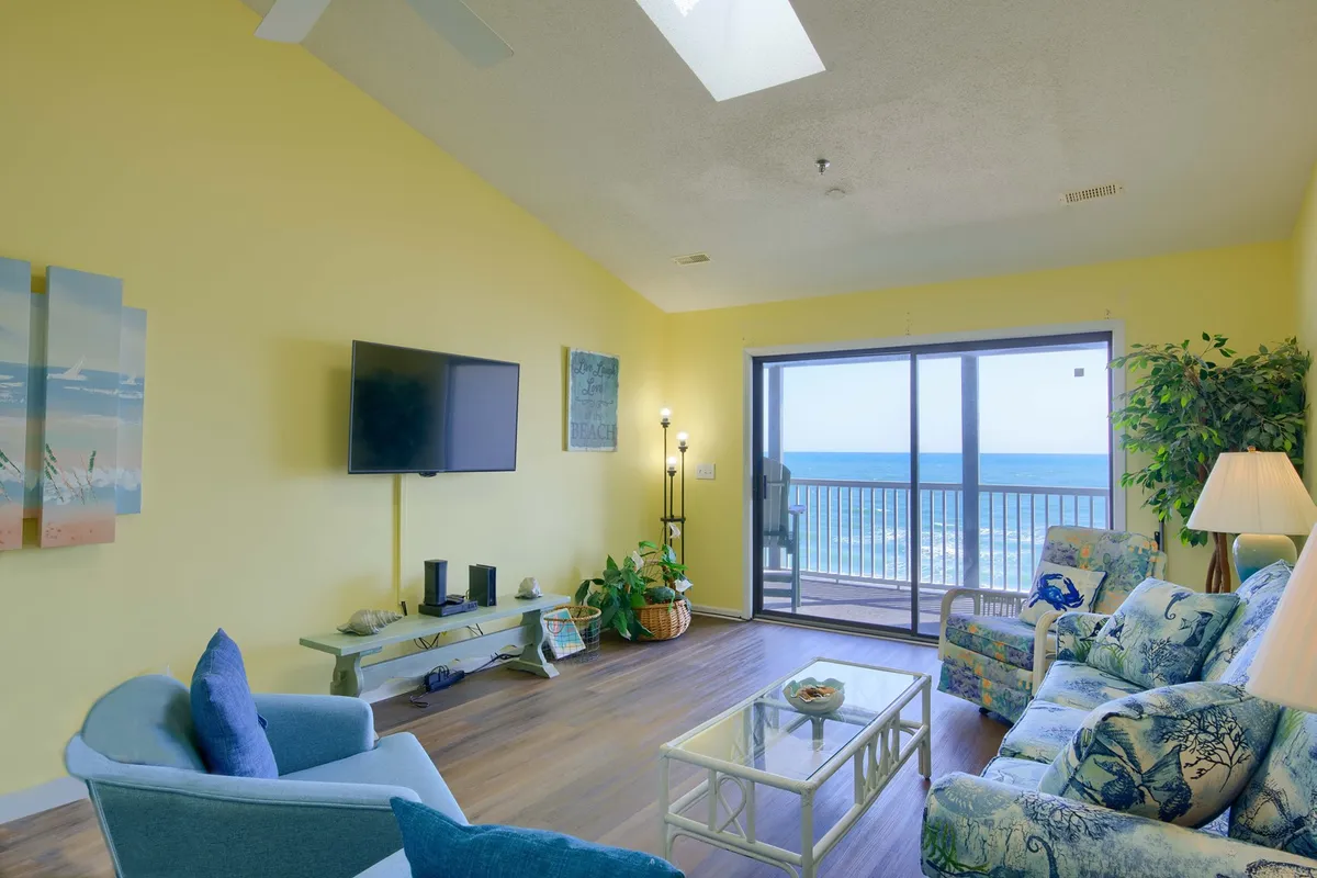 A spacious living room featuring a blue and white floral-patterned sofa facing a large sliding glass door that frames expansive Atlantic ocean views. The room includes a wall-mounted large-screen TV, coastal-themed decorations, and plenty of natural light. The glass doors open out to a balcony, drawing the outdoors in.